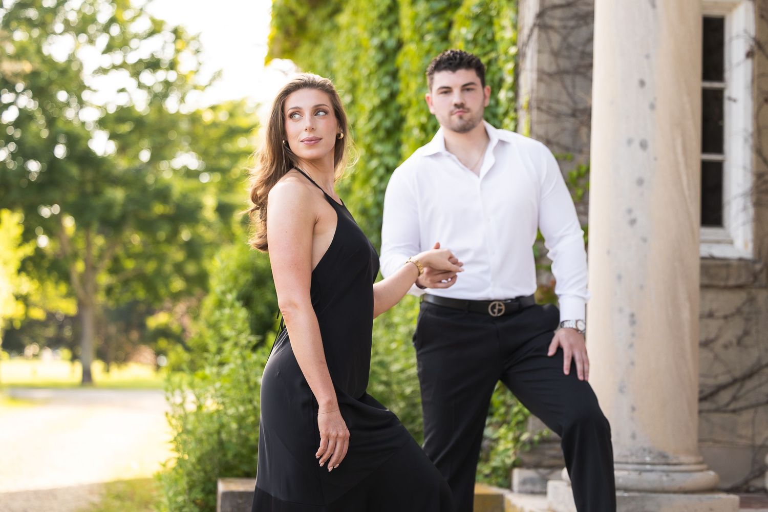Elegant couple in formal attire poses together in a romantic outdoor garden setting during golden hour.