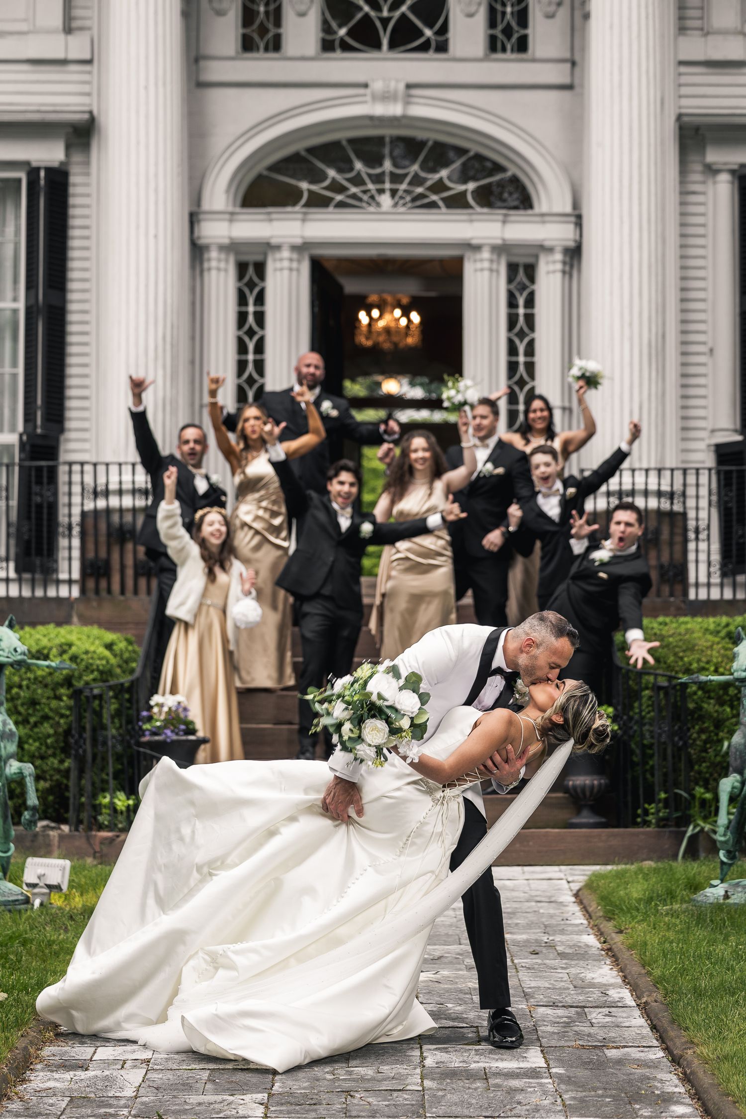 Wedding party performs dramatic dip poses in front of historic white columned building.