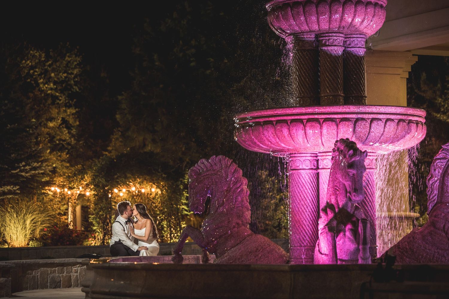 Outdoor wedding venue illuminated with purple uplighting and fountains at night.