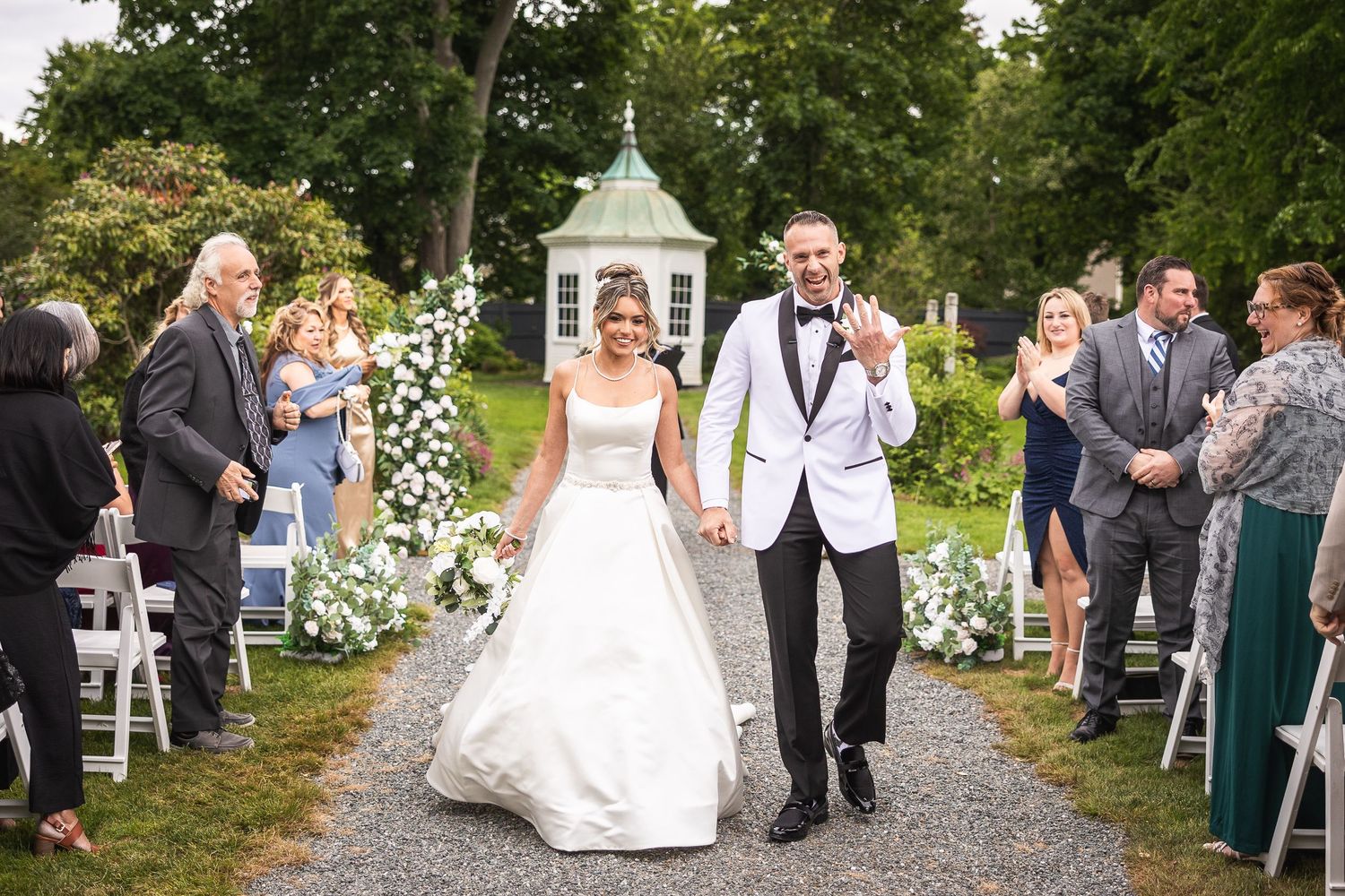 Wedding ceremony guests seated outdoors under trees with white flower decorations.