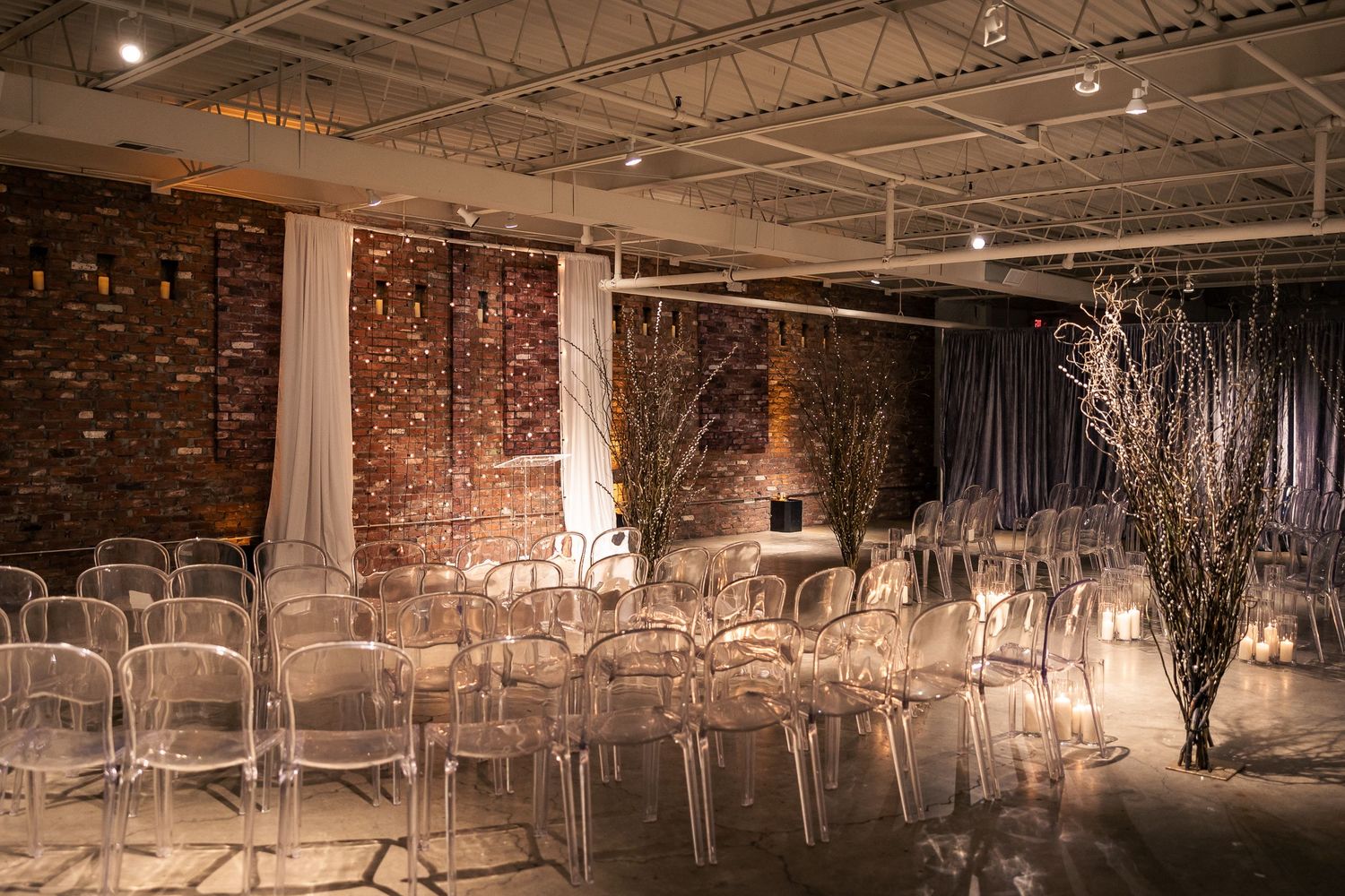 Rustic industrial event space with exposed brick walls, string lights, and clear acrylic chairs arranged in rows.