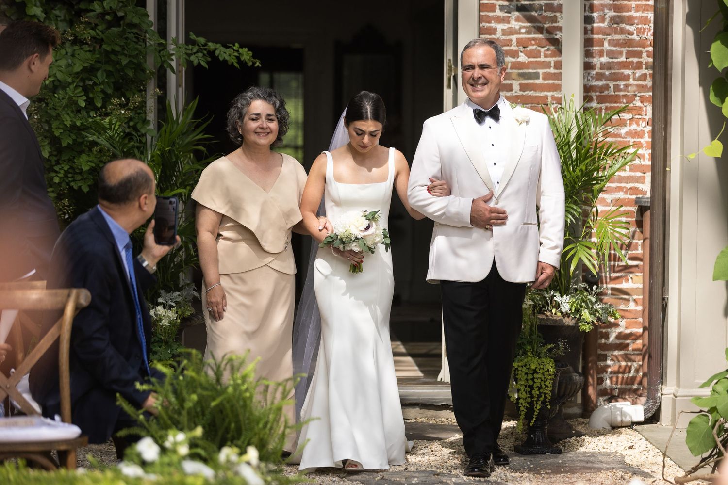 Wedding party gathers in front of brick building during outdoor ceremony.