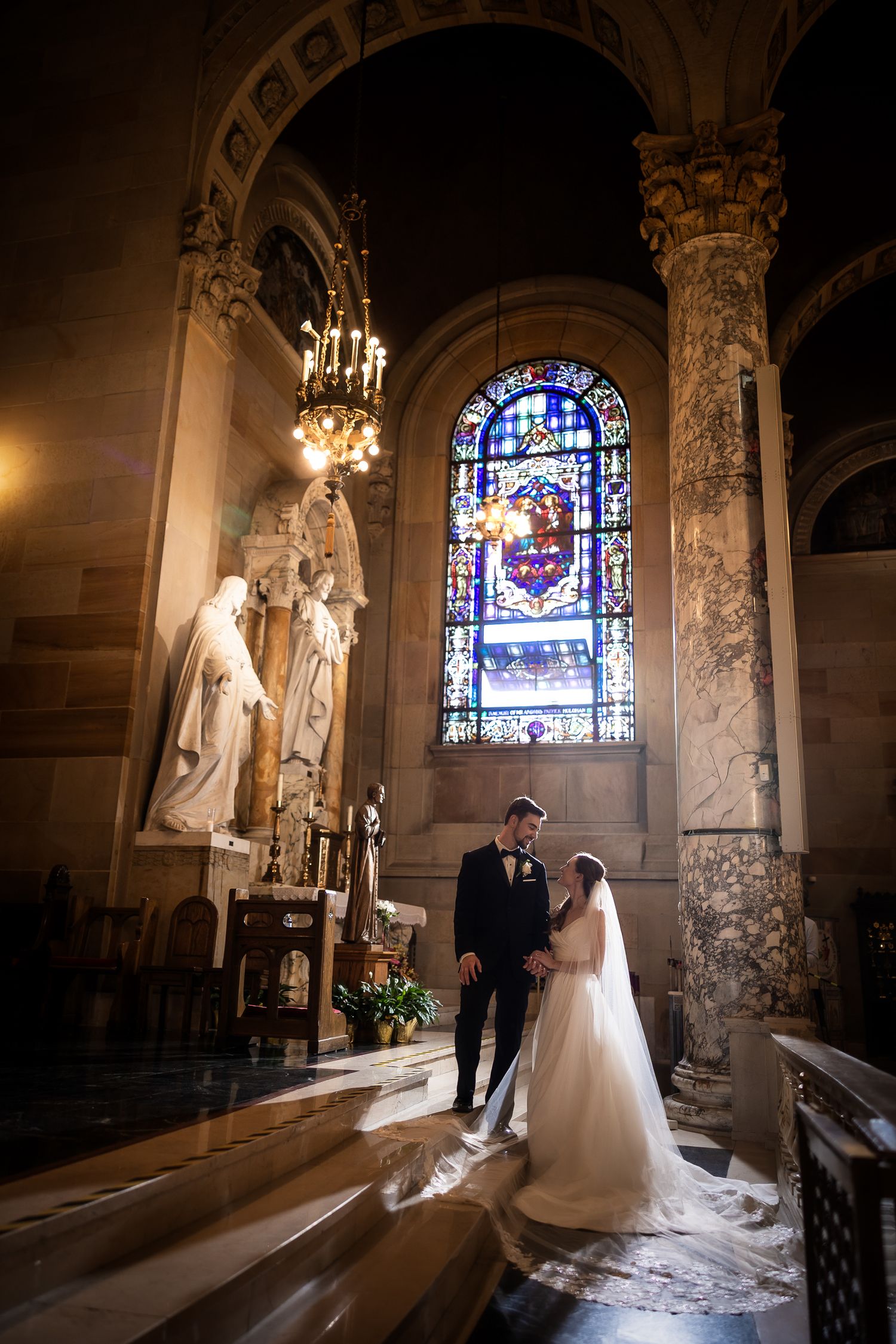 Elegant wedding ceremony in a historic church with stained glass windows and ornate marble columns creates dramatic lighting.