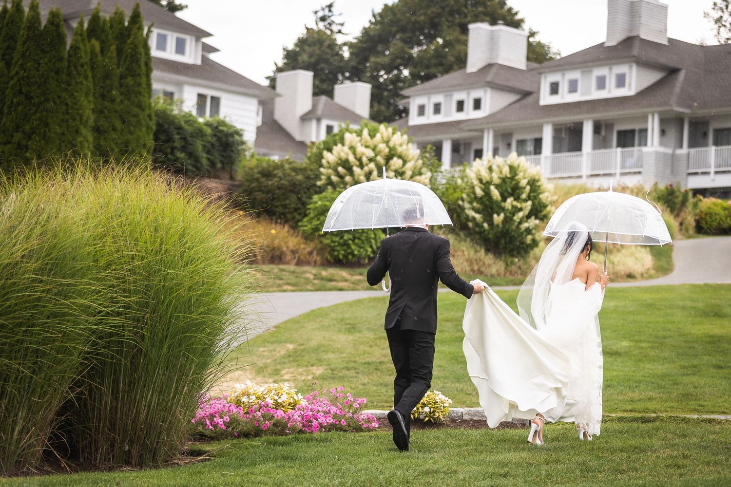 Couple walking with umbrellas on rainy wedding day near white colonial-style building.