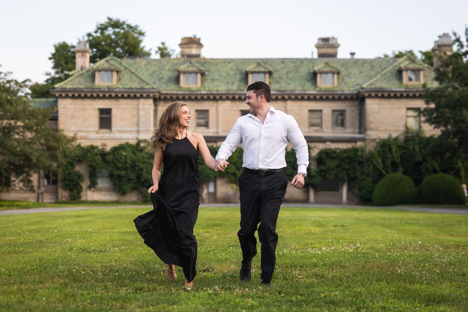 Couple walking together on a grassy hill with historic buildings in the background at sunset.