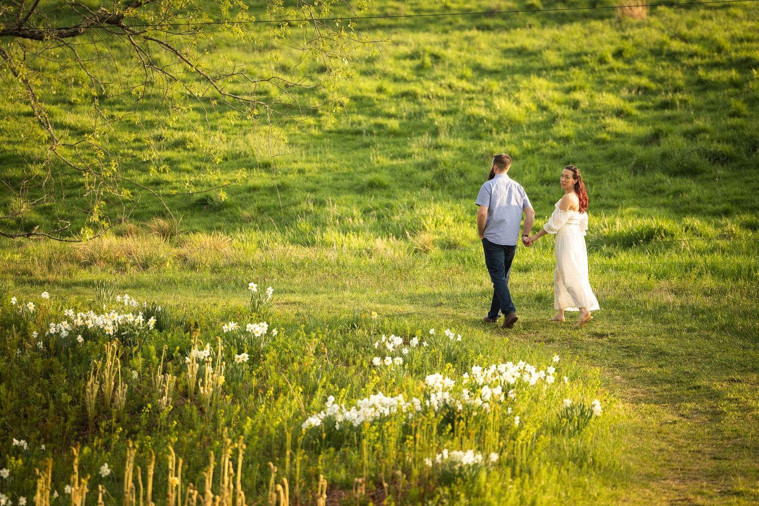 Two people walk together across a sunlit meadow filled with wildflowers on a warm summer afternoon.