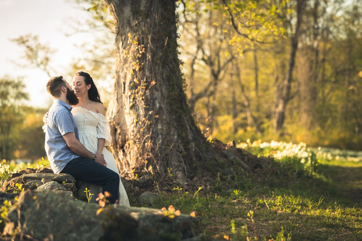 A couple sits together on fallen autumn leaves in a sunlit forest during golden hour.
