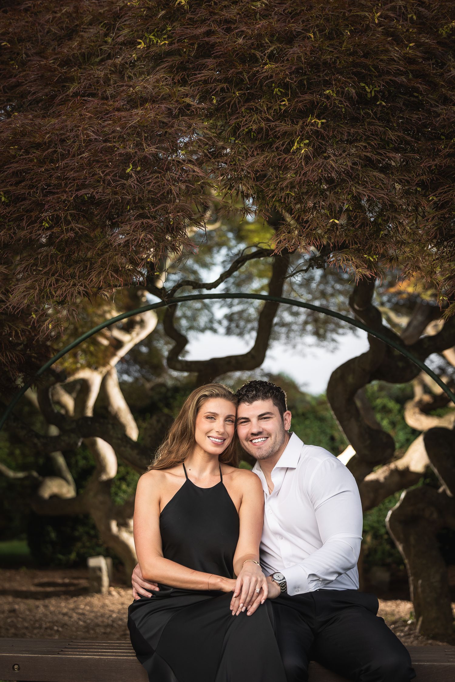 A romantic outdoor portrait under twisted tree branches creating a natural archway in a park setting.