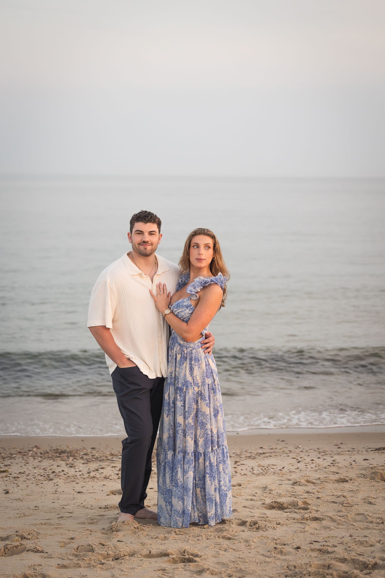 A couple stands together on a misty beach at sunset wearing coordinated blue and white outfits.