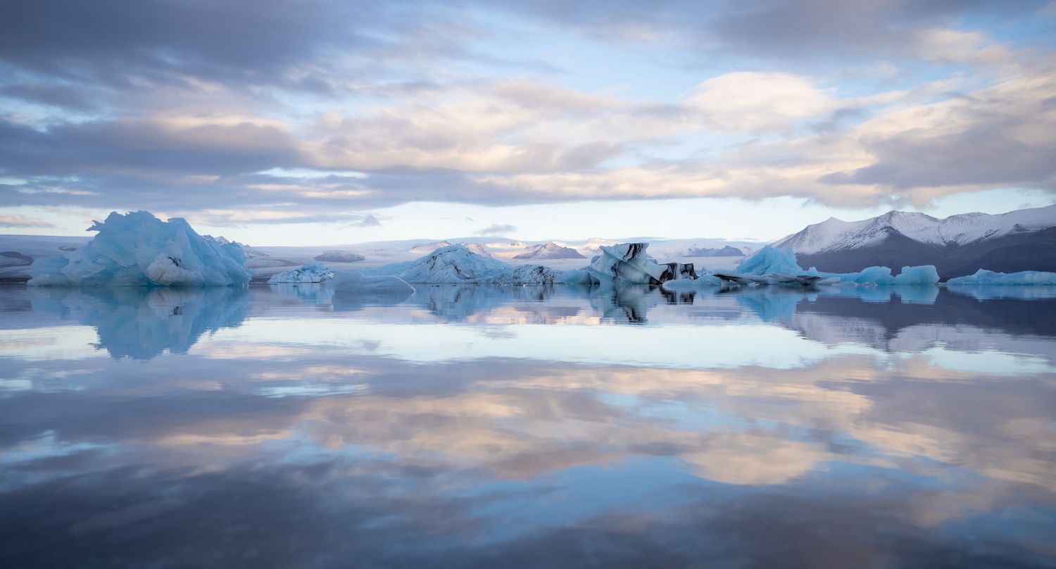 Stunning reflection of icebergs and mountains on still glacial lake under cloudy sky.