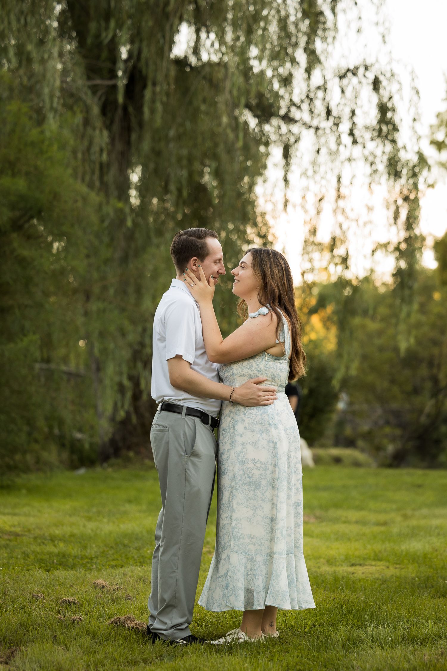A couple embraces and gazes at each other lovingly in a lush green park beneath a weeping willow tree at golden hour.