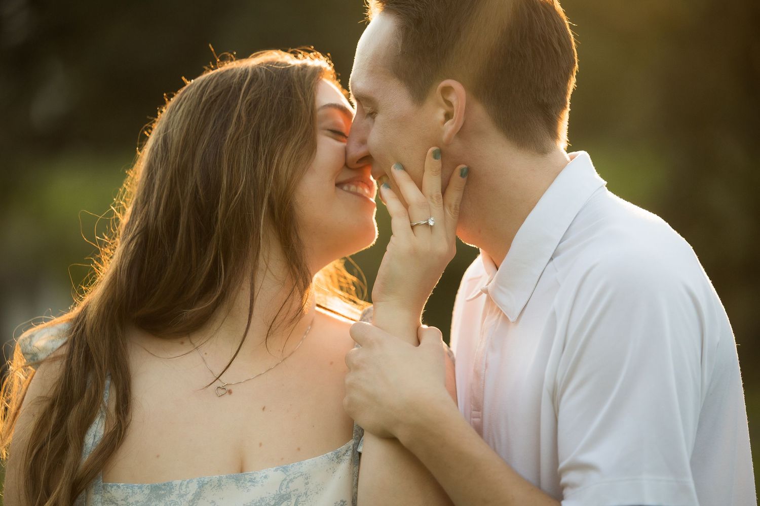 Couple sharing a romantic almost-kiss moment outdoors, bathed in warm golden sunlight, woman wearing engagement ring.