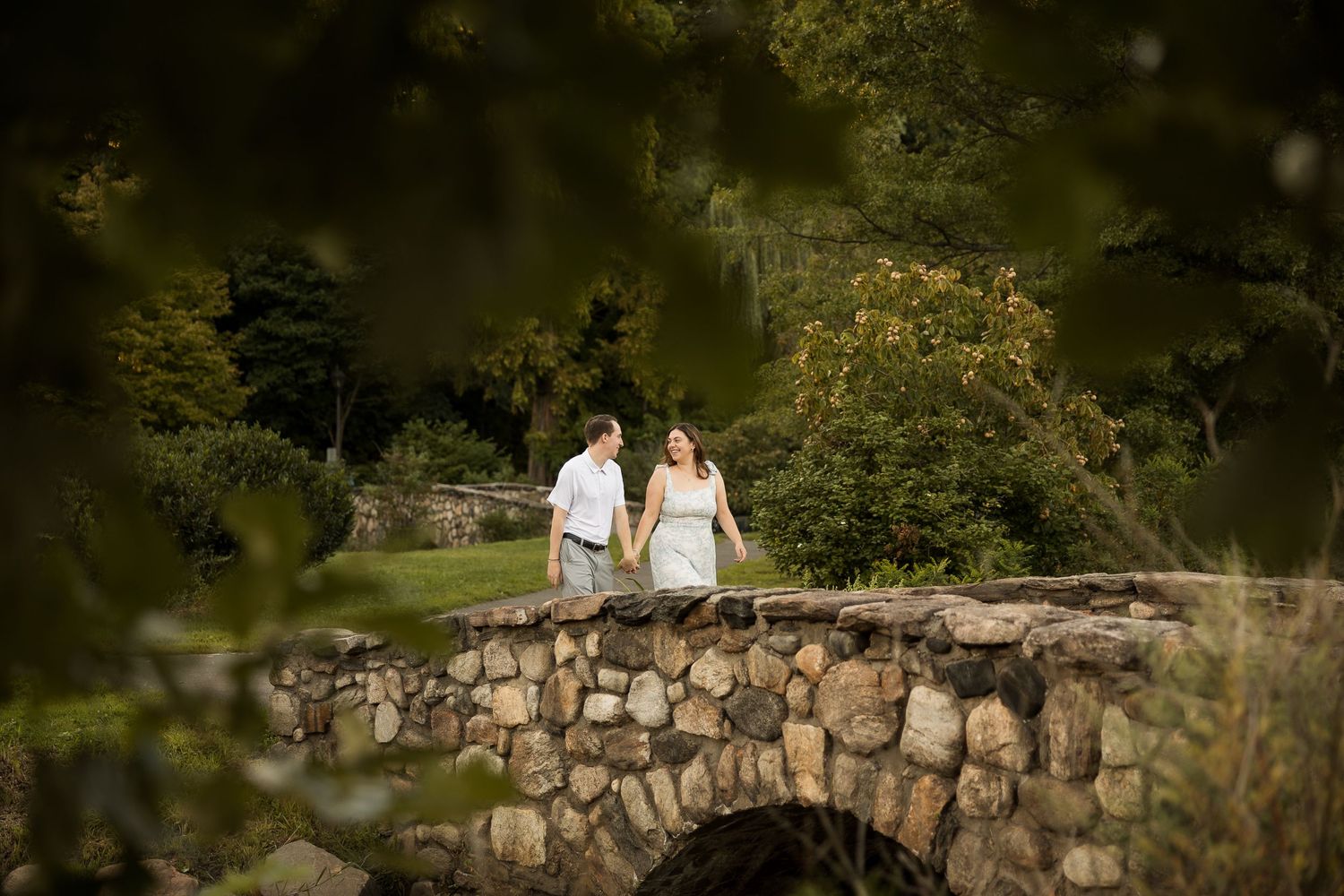 Couple in white walks hand-in-hand on a stone arch bridge surrounded by lush green trees in a garden setting.