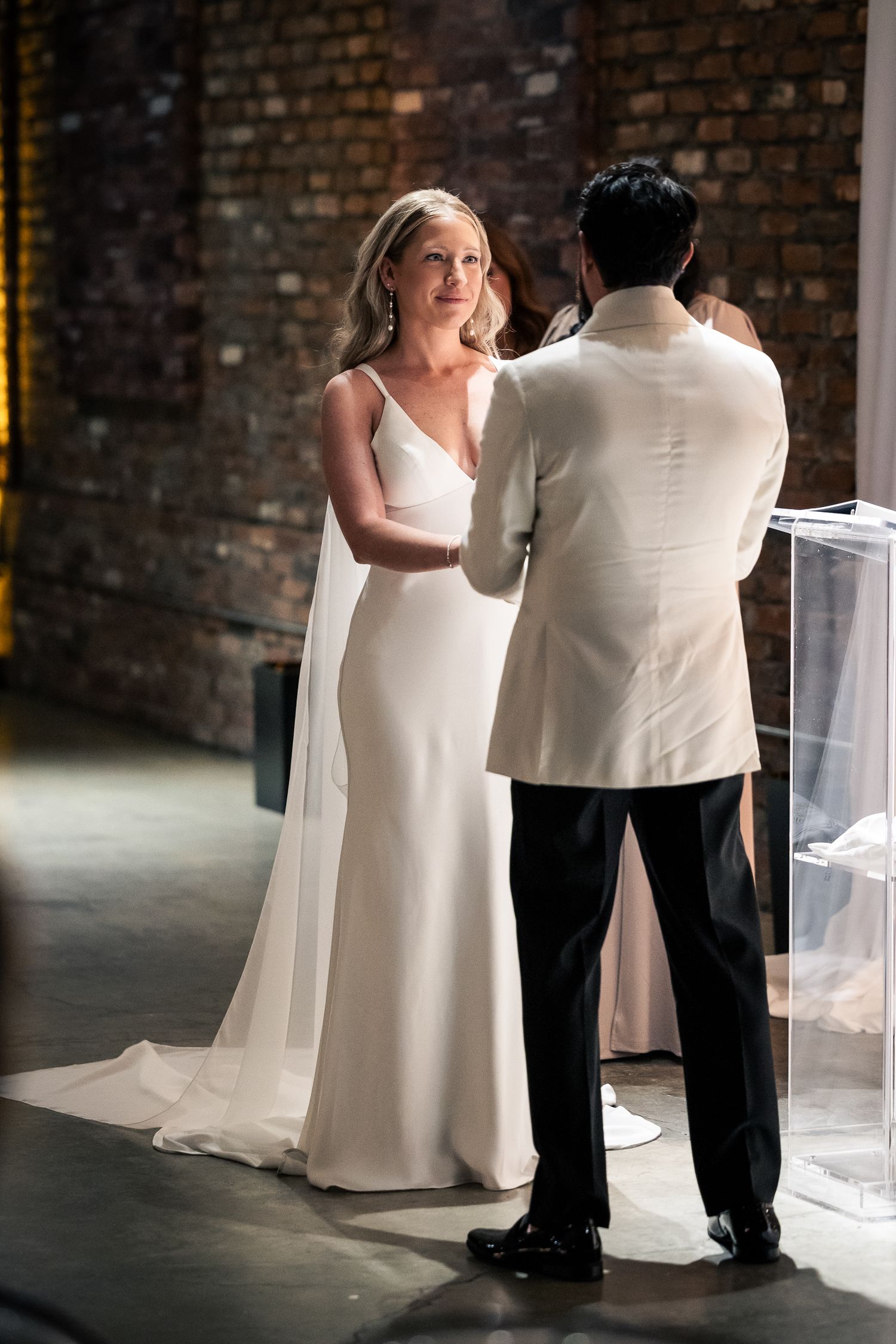 A couple in white and black formal attire dance together in a brick-walled venue with dramatic lighting.