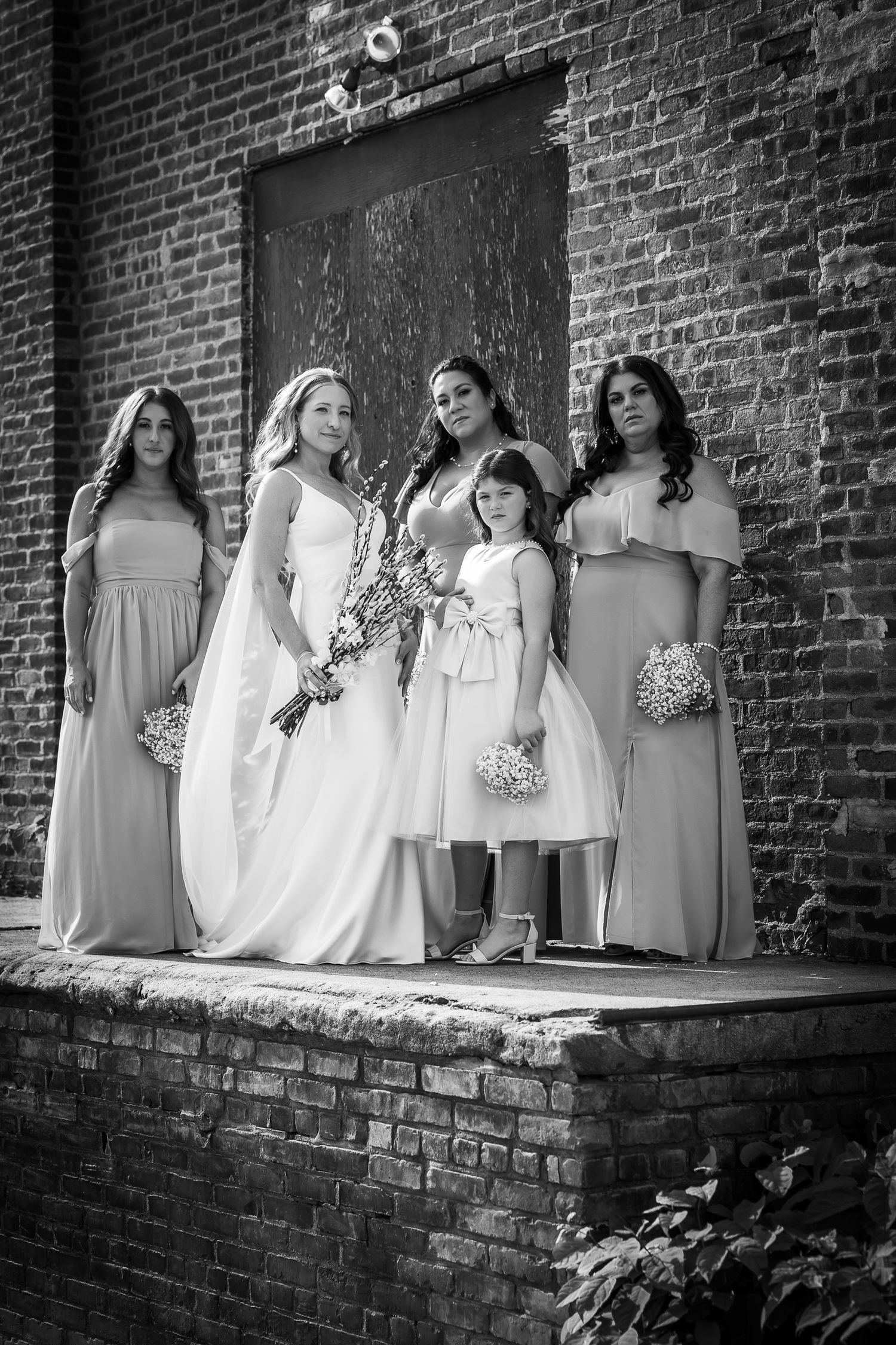 Black and white wedding party poses on rustic brick wall ledge in elegant dresses with bouquets.