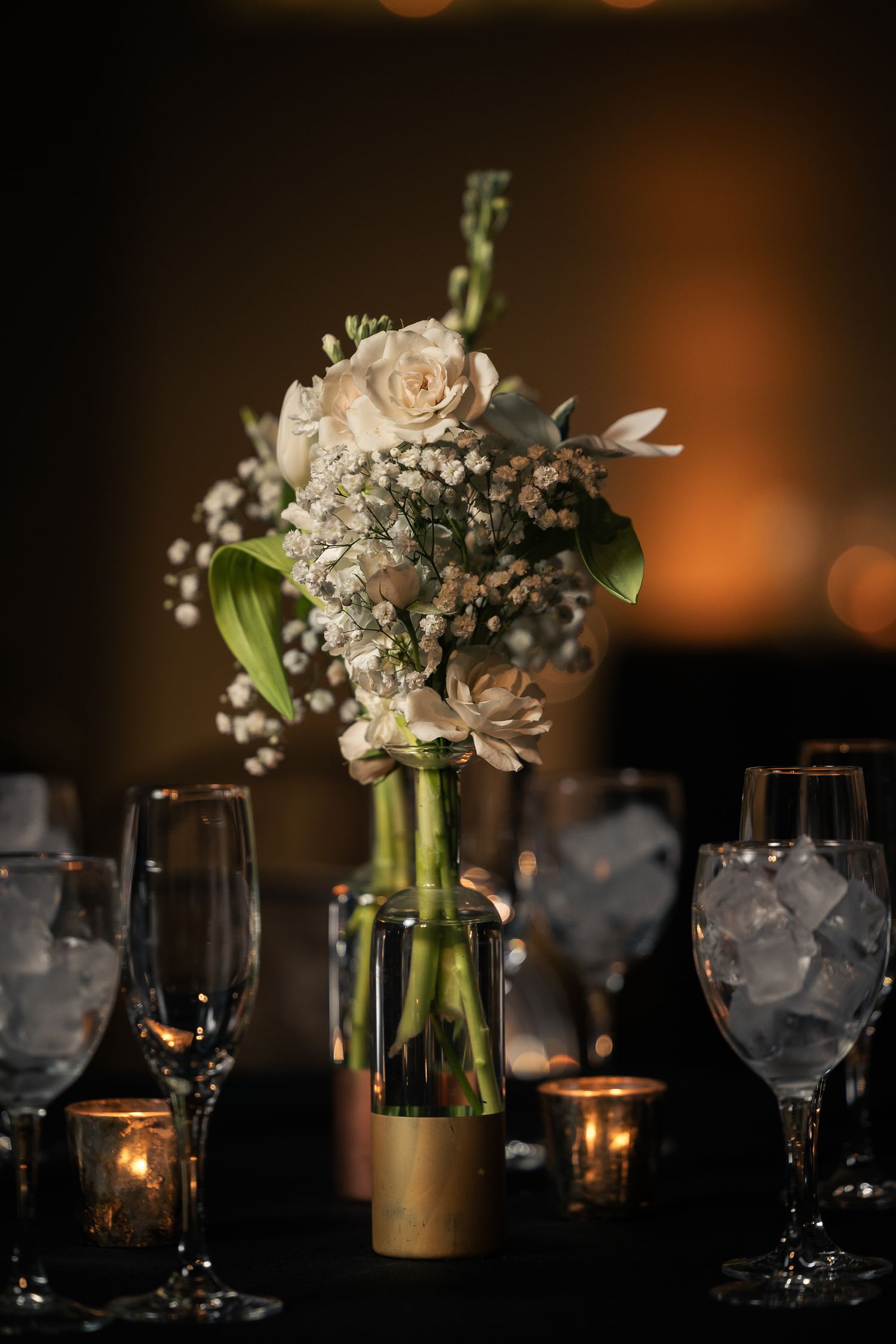Elegant white floral centerpieces with roses and baby's breath in glass vases illuminate a dark table with candlelight and wine glasses.
