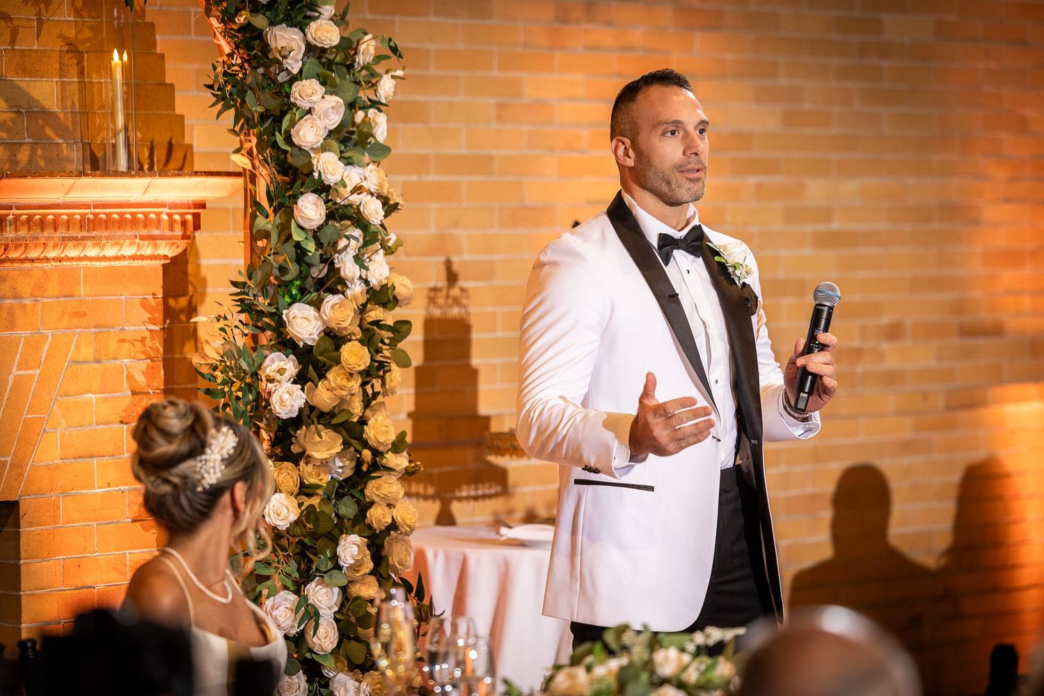Wedding party poses against warm-lit brick wall decorated with white floral arrangements.