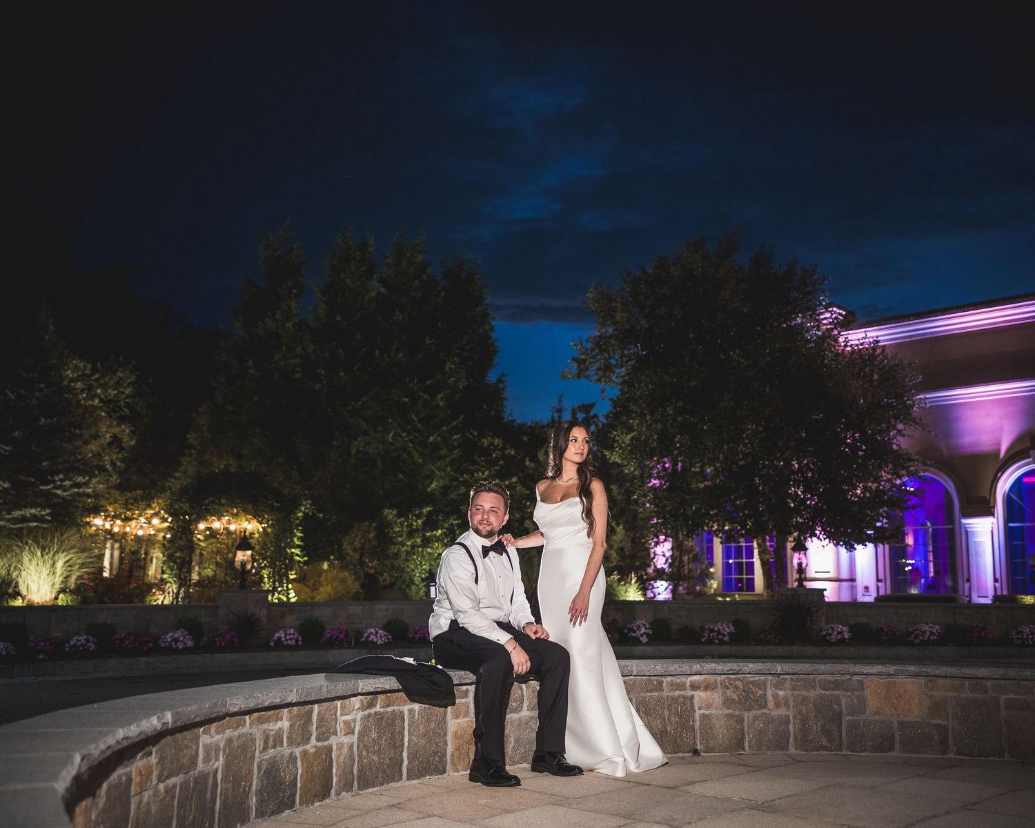 Evening wedding portraits on a stone terrace with purple uplighting and trees in background.