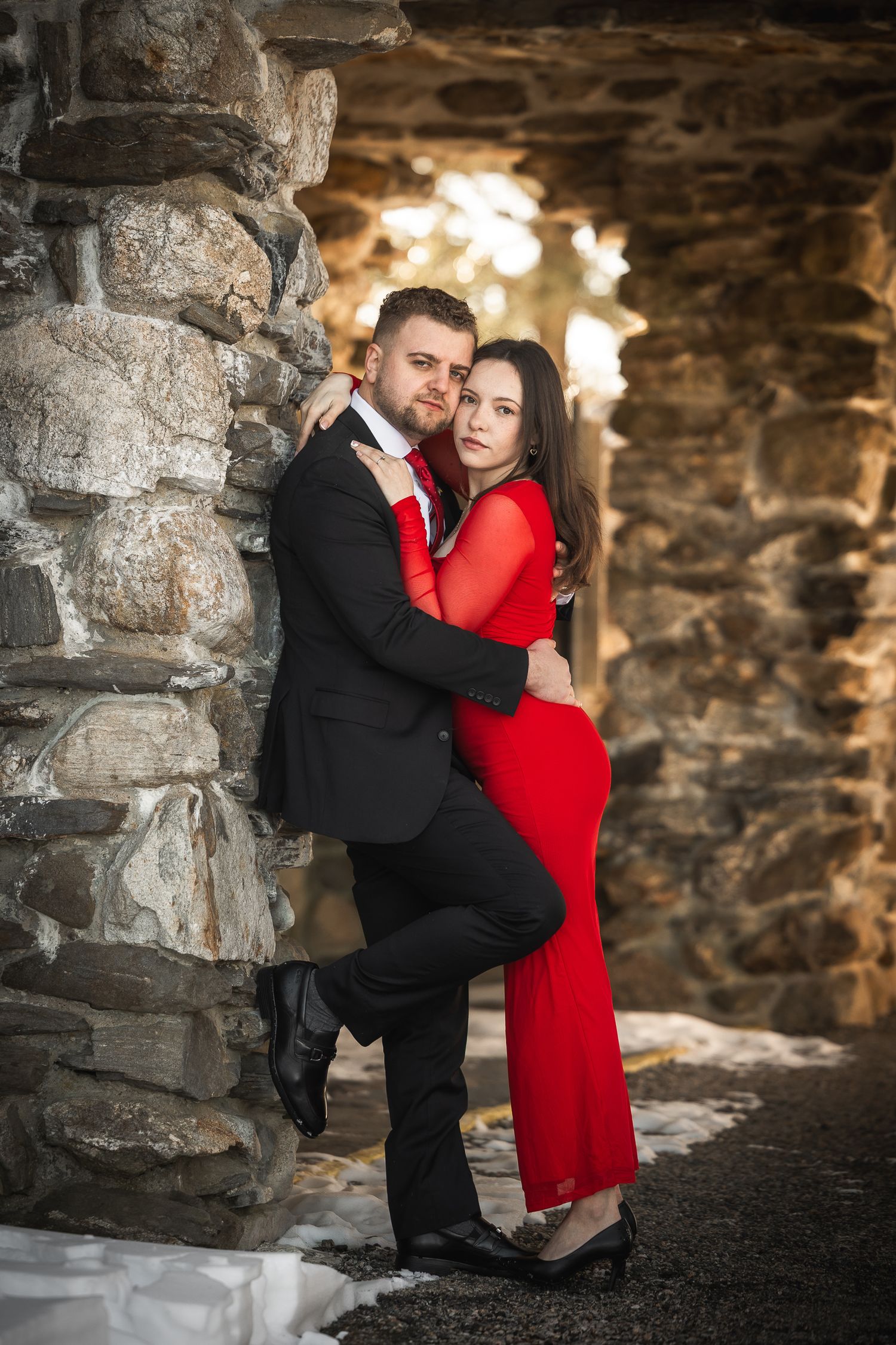 A romantic couple embraces against a stone wall background, with the person in red wearing a long dress and other in black suit.