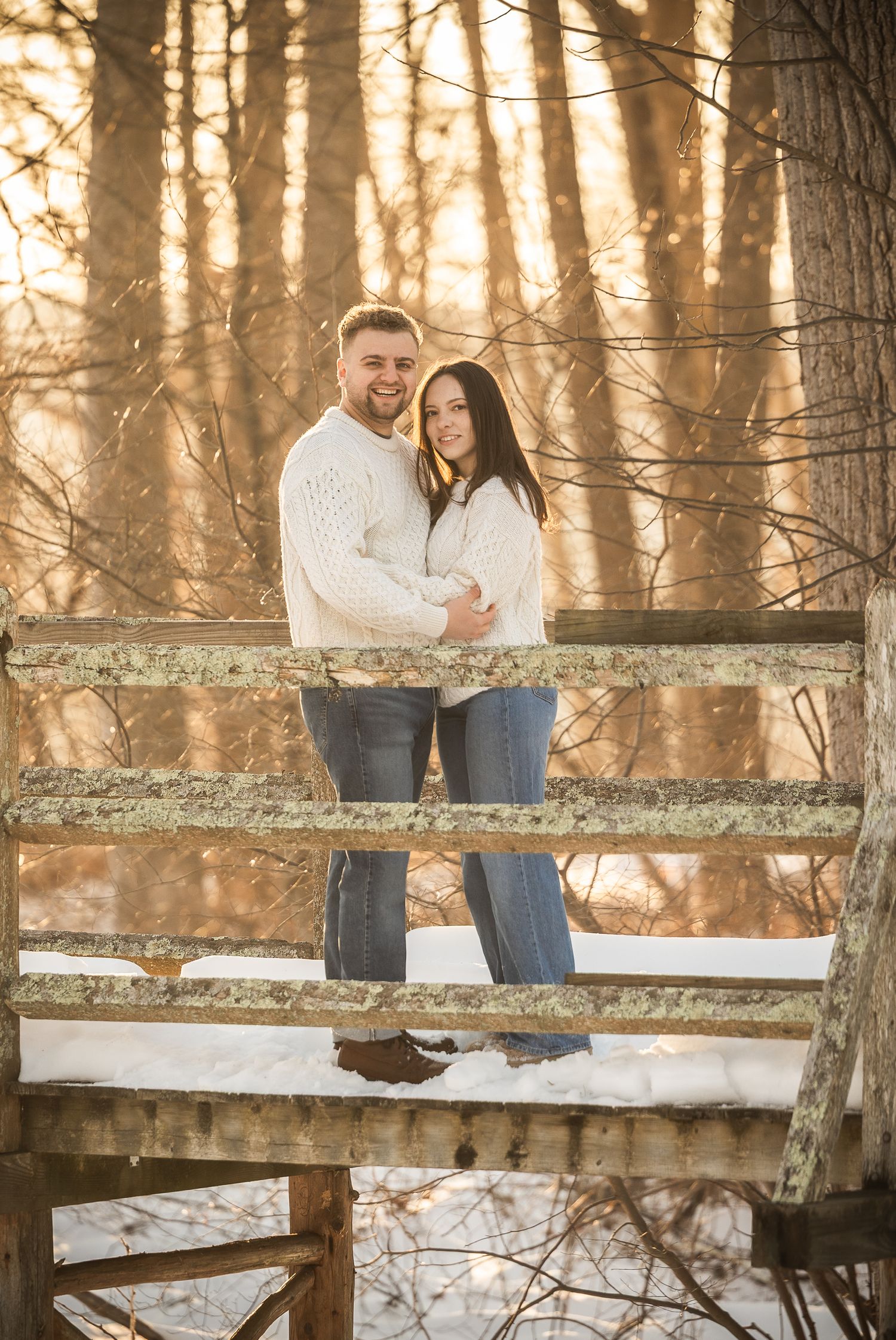 A couple sits together on a wooden fence in a snowy forest during golden sunset light.