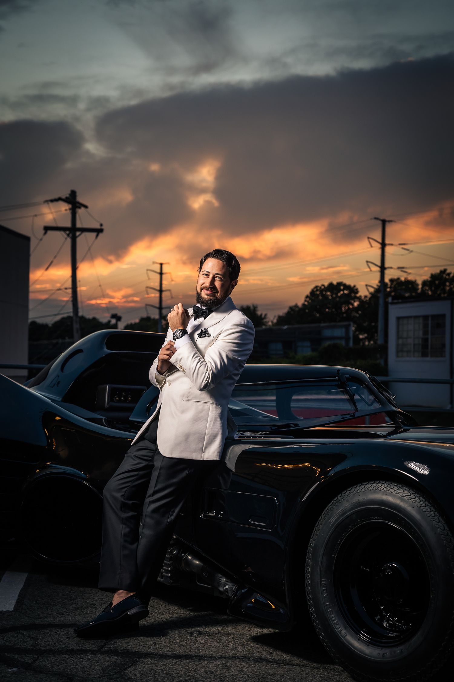 A dramatic sunset silhouettes utility poles and a vintage black car in a moody urban scene.