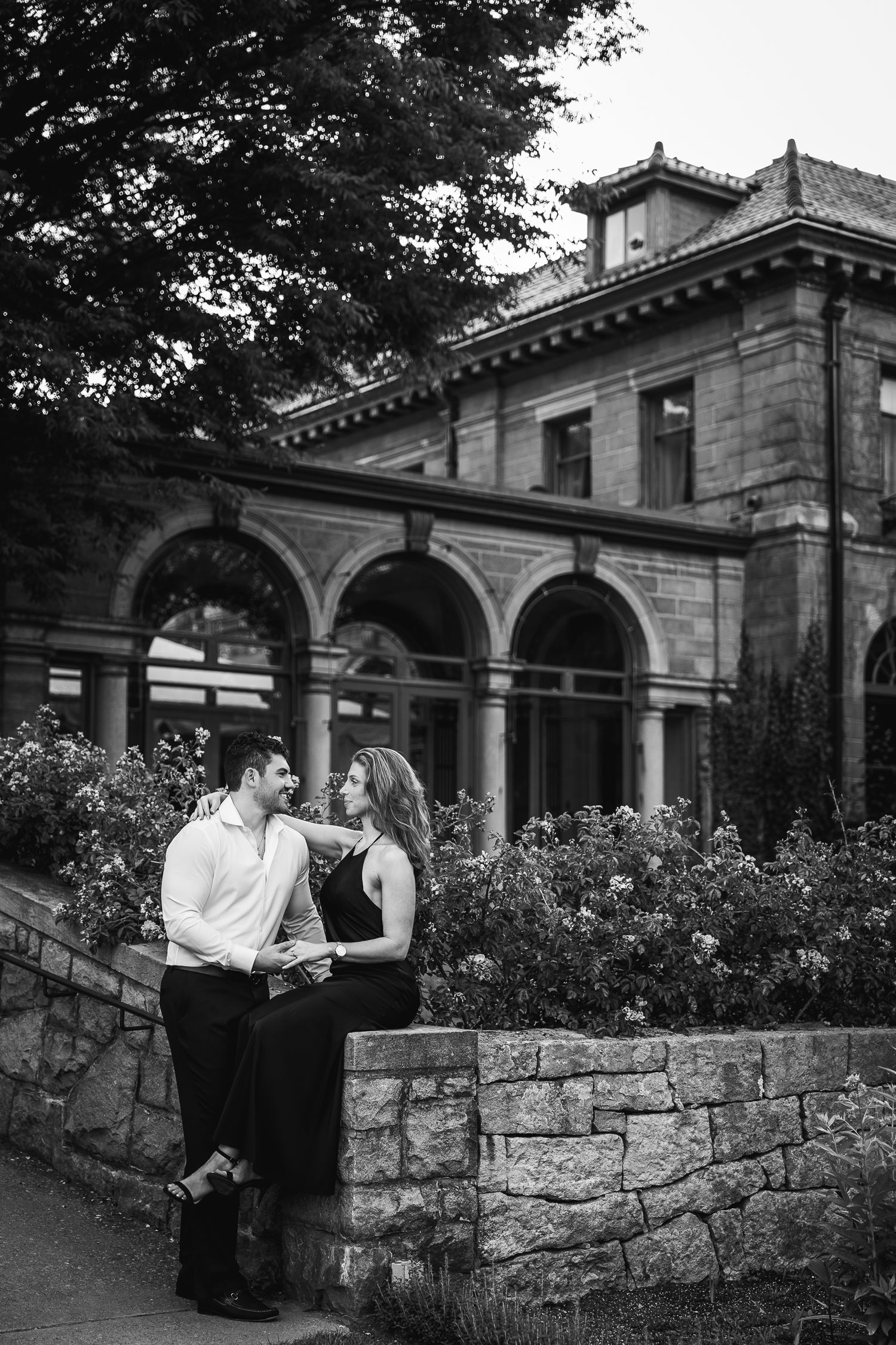 A romantic couple shares tender moments along a stone wall with arched architecture in black and white photography.