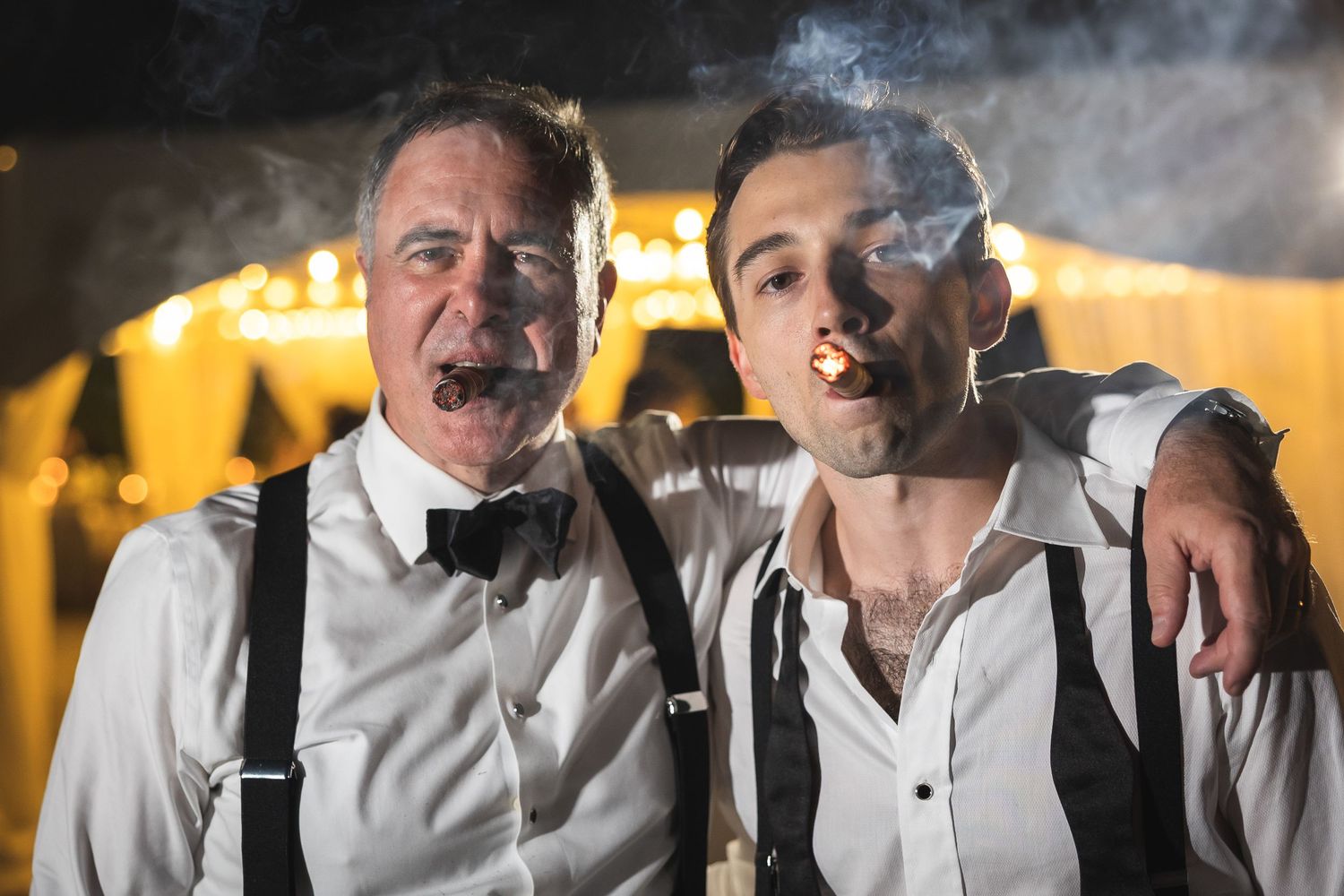 Groom with father-in-law smoking cigars at an evening wedding celebration.