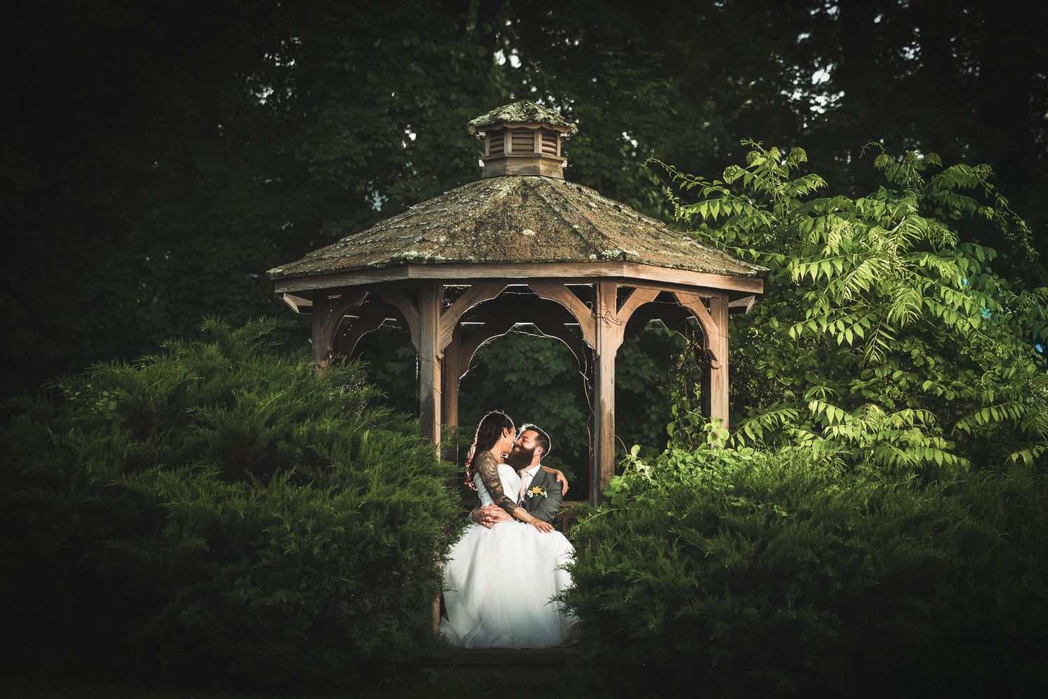 A couple shares a romantic kiss under a wooden gazebo surrounded by lush green garden scenery.