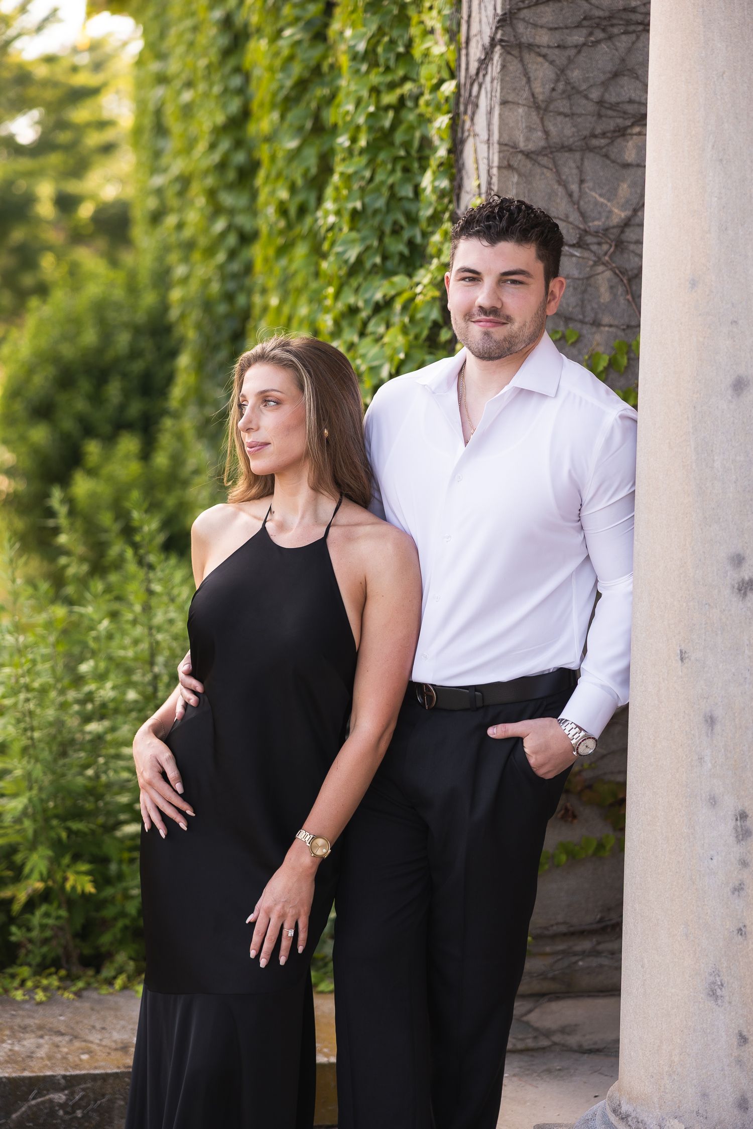 A couple in contrasting black and white outfits pose together in an outdoor setting with green foliage in the background.