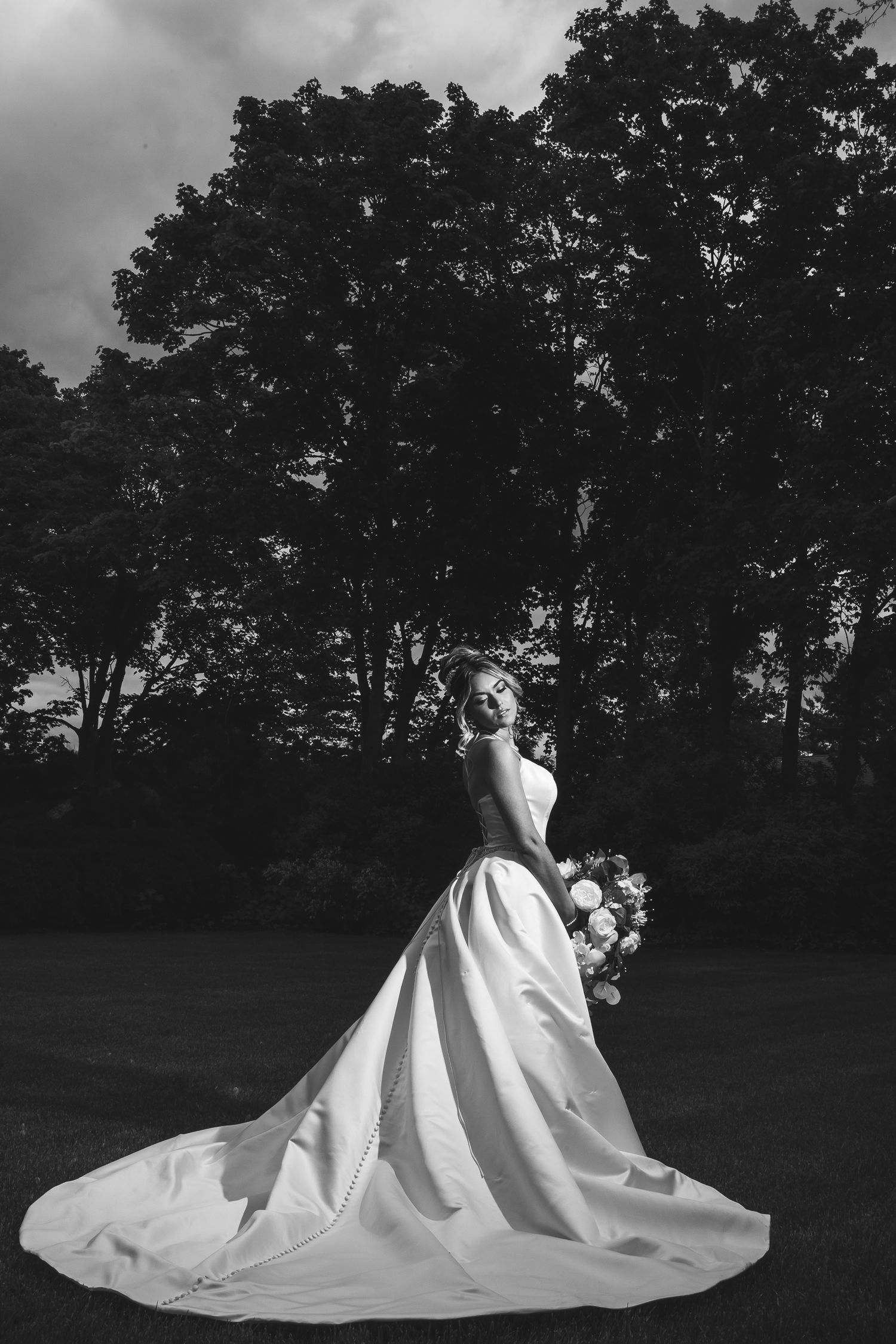 A flowing white wedding dress train spreads across a dark lawn at dusk with trees in the background.