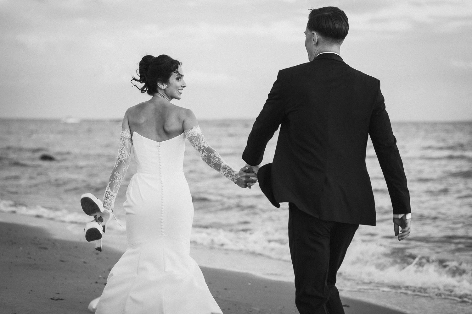 Black and white photo of newlyweds walking hand in hand along a beach at sunset.