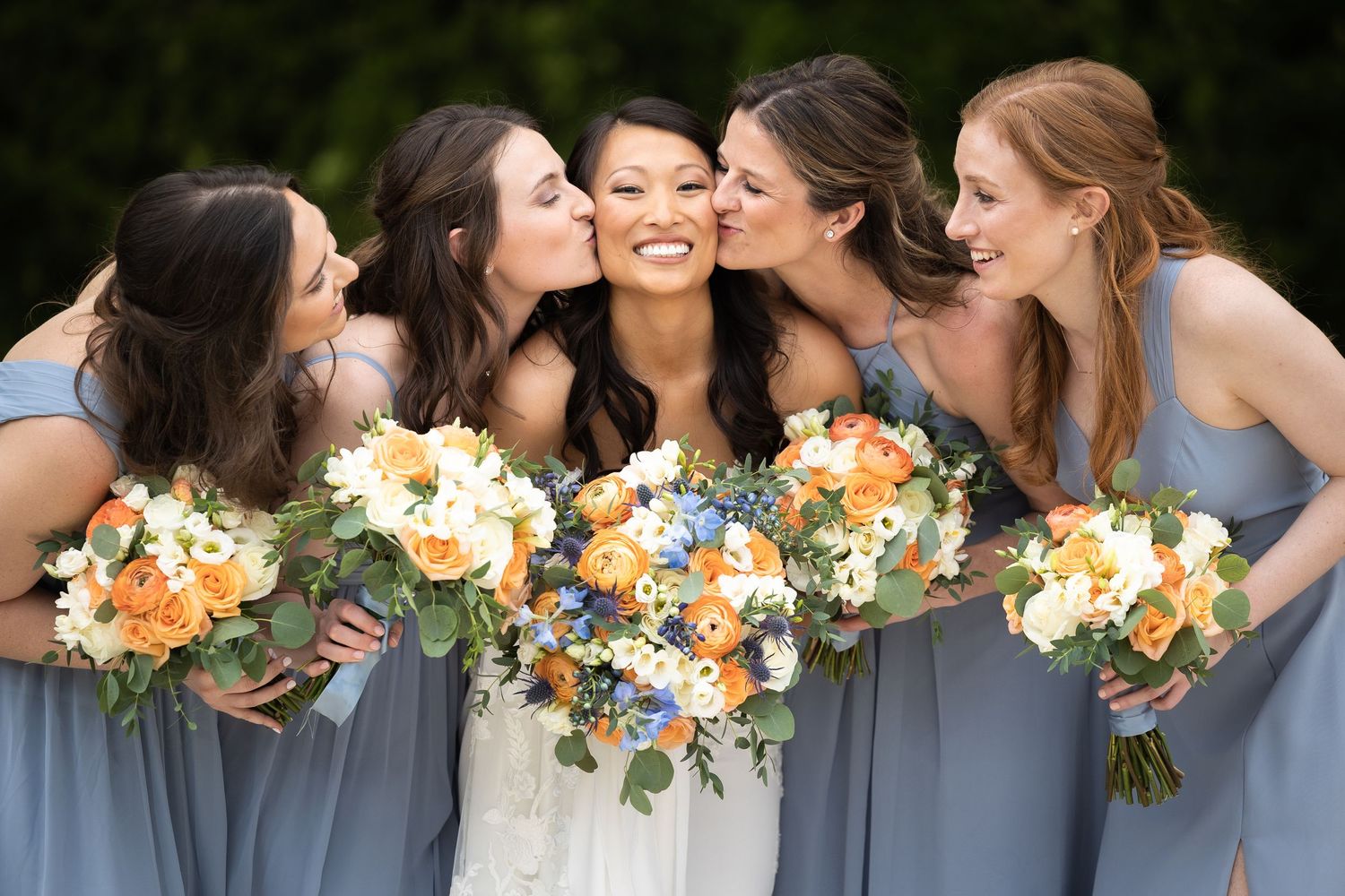 Bridesmaids in blue dresses hold peach and white bouquets while posing together.