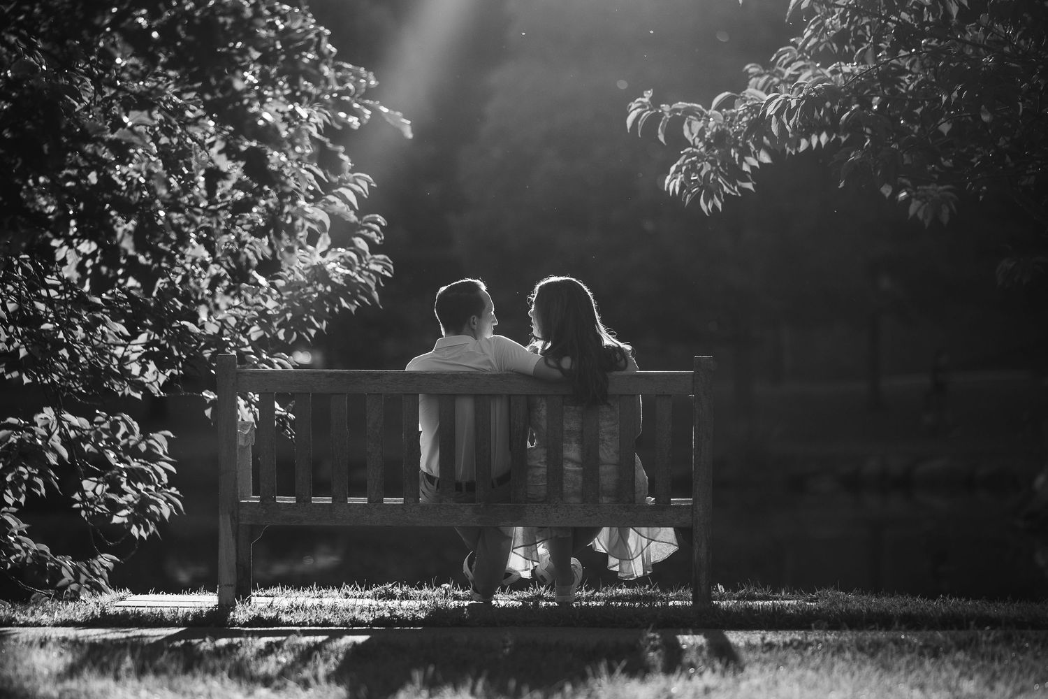 Black and white photo of a couple sitting together on a wooden bench in a sunlit park surrounded by lush trees.