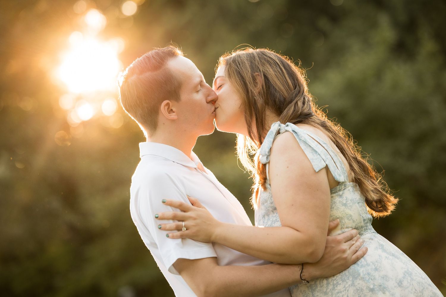 Couple sharing a romantic kiss outdoors during golden hour sunset with warm glowing light and green trees.