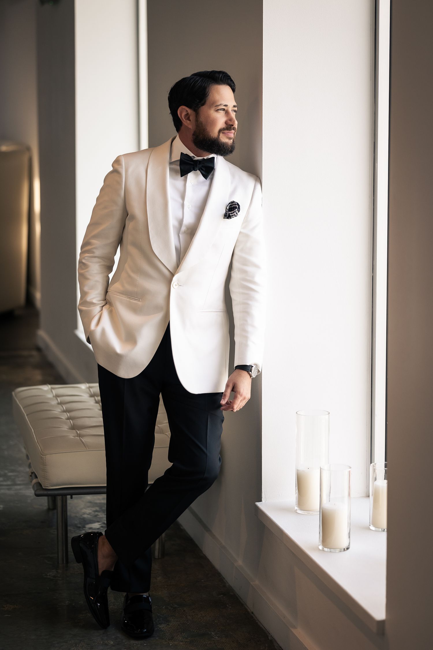 A figure in a white dinner jacket and black pants stands contemplatively by a window with candles in an elegant setting.