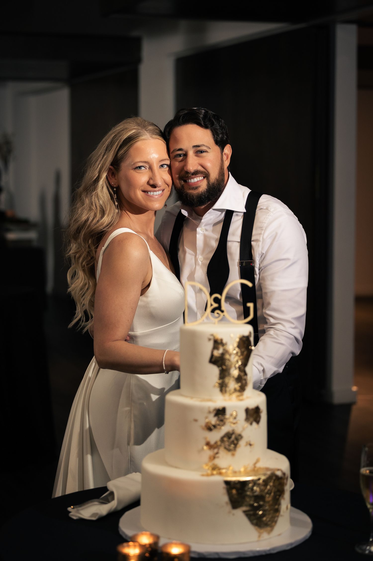 A couple shares a joyful moment during their wedding cake cutting ceremony in a dimly lit reception venue.