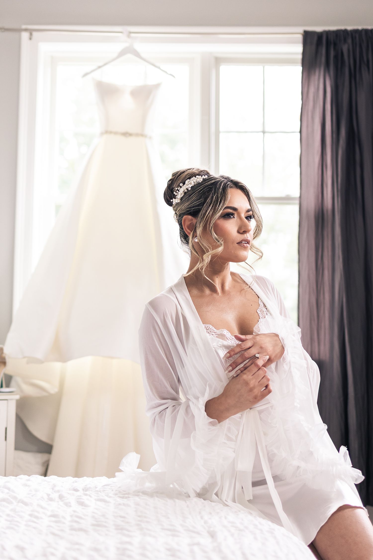 Bridal preparation scene with white dress hanging in sunlit room with dark curtains.