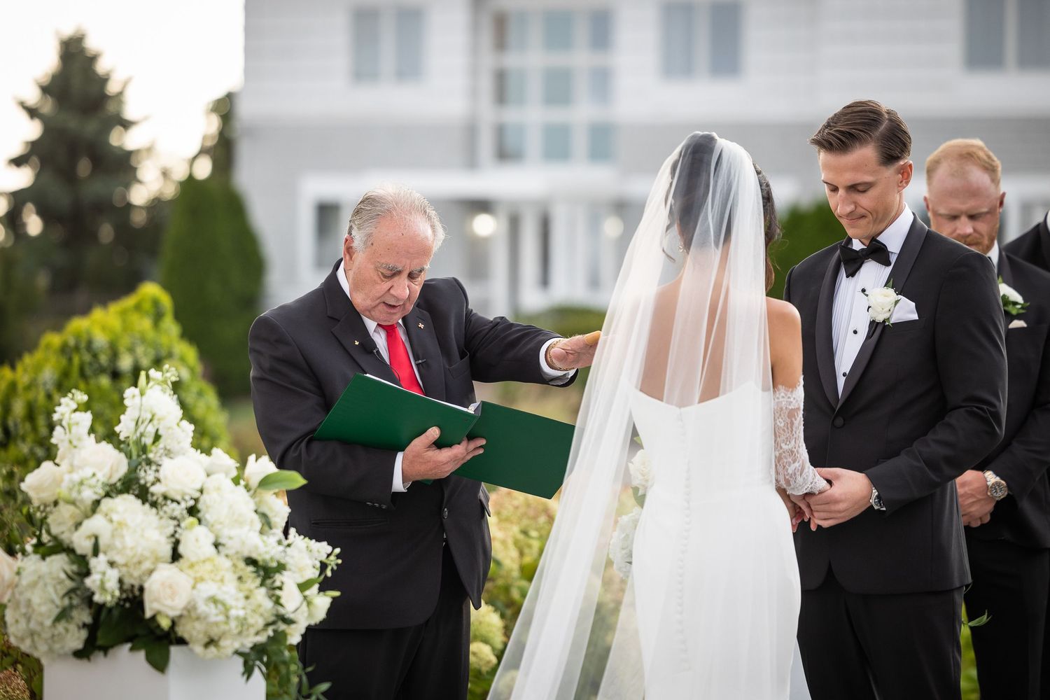 Wedding ceremony taking place outdoors with an officiant reading from a book beside floral arrangements.