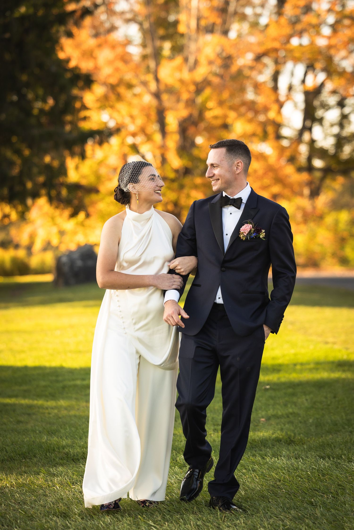 A couple in wedding attire walks arm in arm through a park with vibrant autumn foliage in the background.