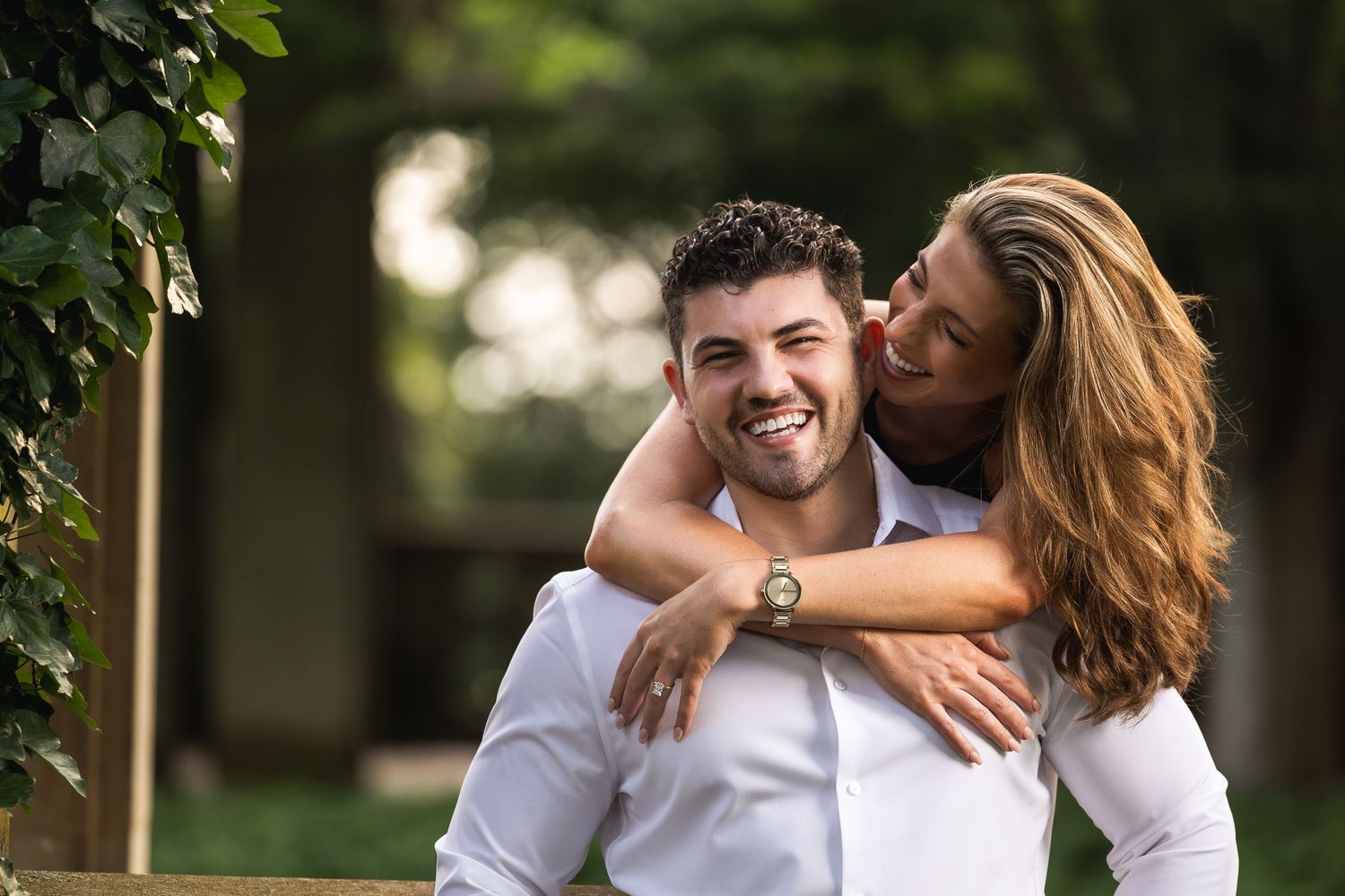 A romantic couple embraces and laughs together in a sunny outdoor garden setting.