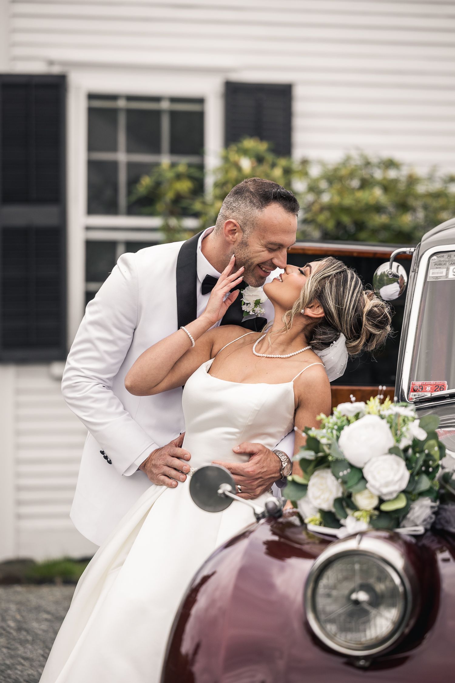 A romantic moment captured beside a vintage burgundy car at a wedding.