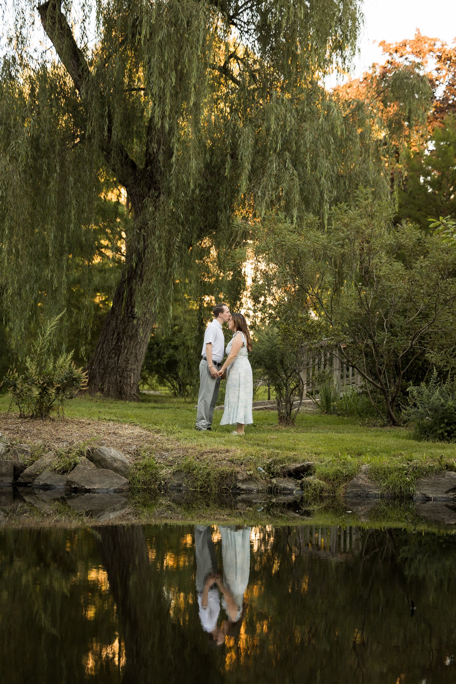 Couple sharing a romantic moment by a reflective pond under a weeping willow tree in a lush garden setting.