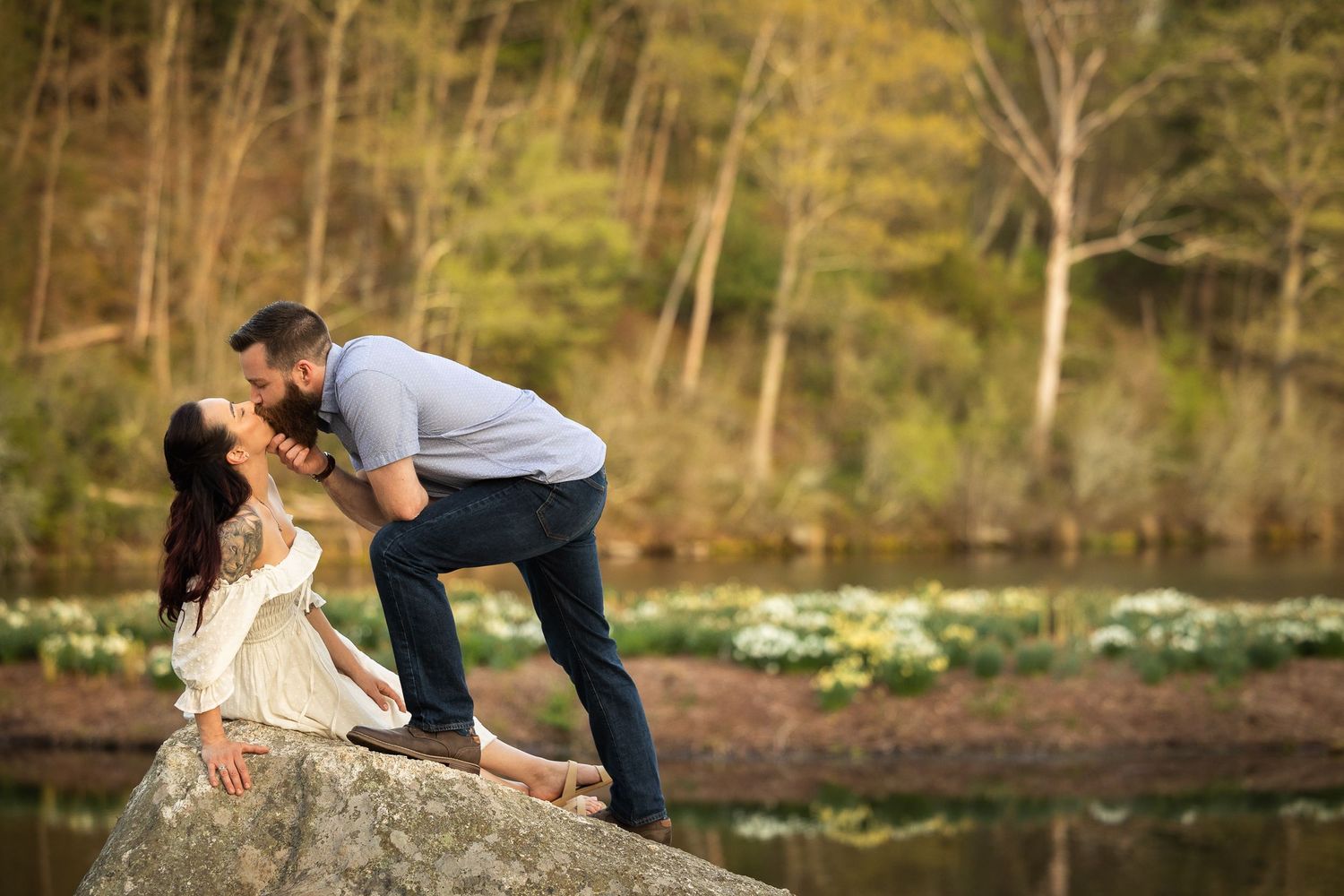 A couple shares a romantic moment by a scenic river with trees and rocks in the background.