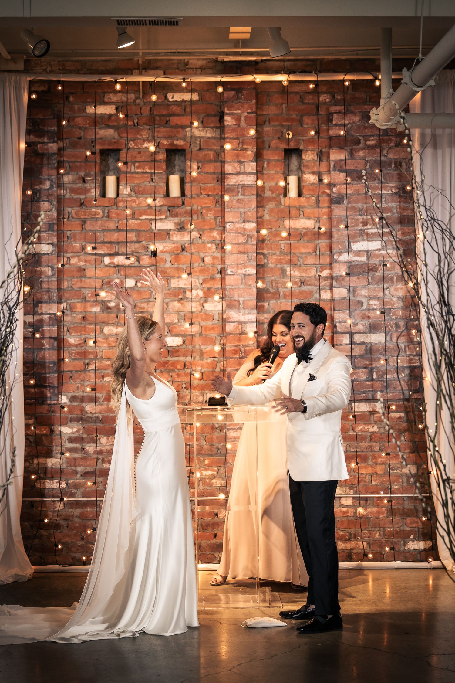 Wedding party dancing and celebrating in front of rustic exposed brick wall with romantic lighting and decor.