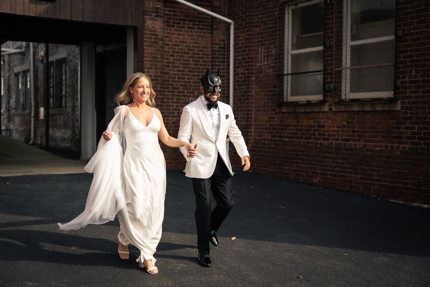 A couple in wedding attire runs playfully along a brick building exterior during their photo session.