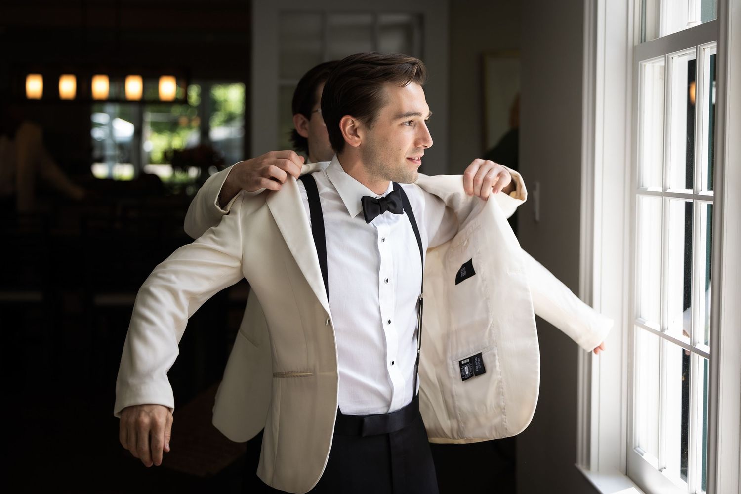 Person in white formal attire standing by bright windows in an elegant room.