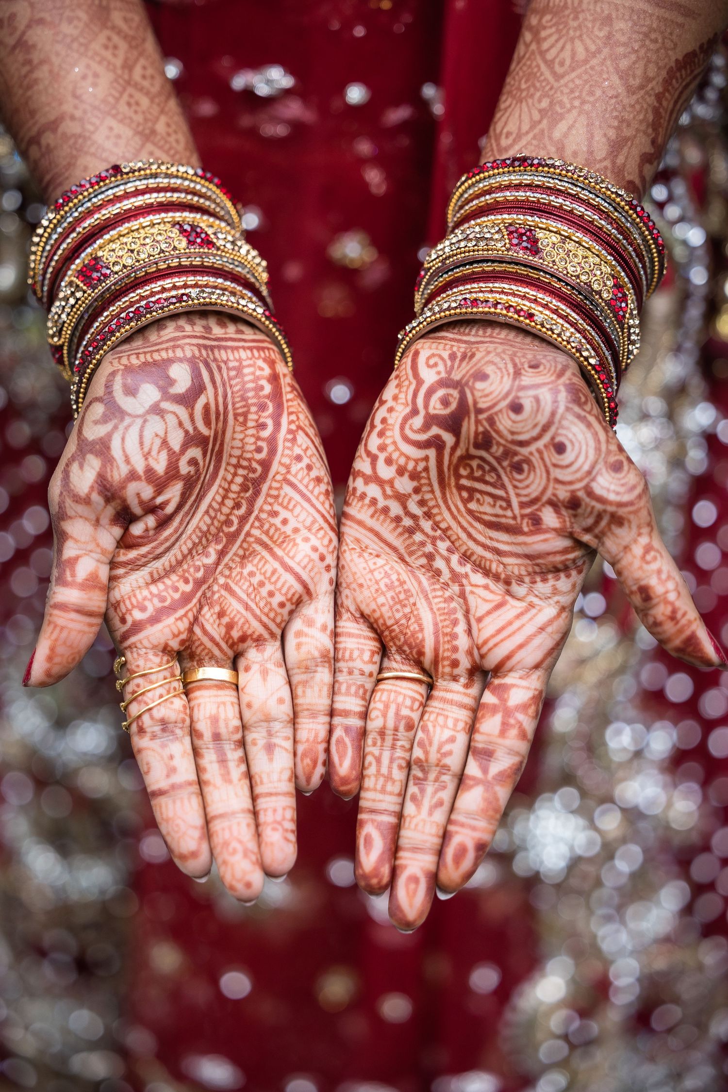Detailed henna mehndi designs on palms with traditional bangles against a red background with bokeh effect.