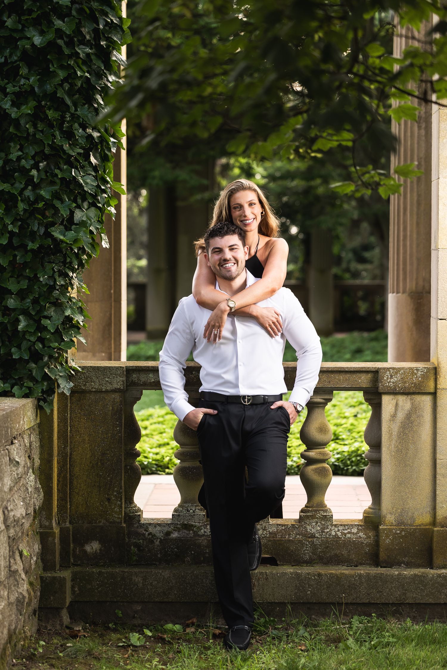 A couple shares a romantic moment on a stone balcony surrounded by lush greenery in an outdoor garden setting.