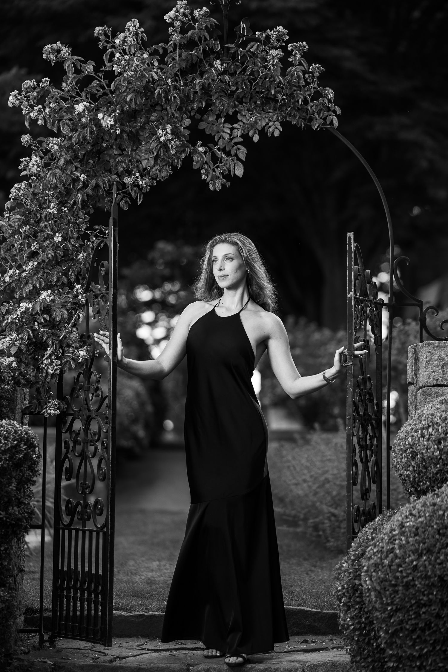 A woman in a flowing black dress dances gracefully near an ornate garden gate in a moody black and white photograph.