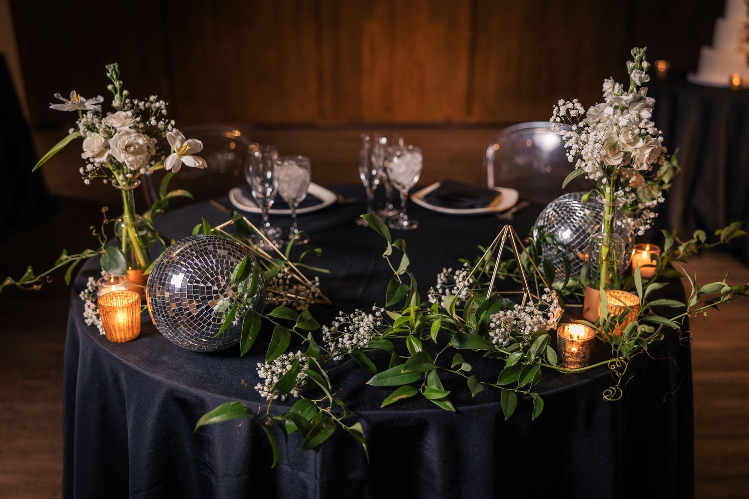Elegant wedding reception table with white floral arrangements, glowing lanterns, and navy blue linens in a rustic venue.